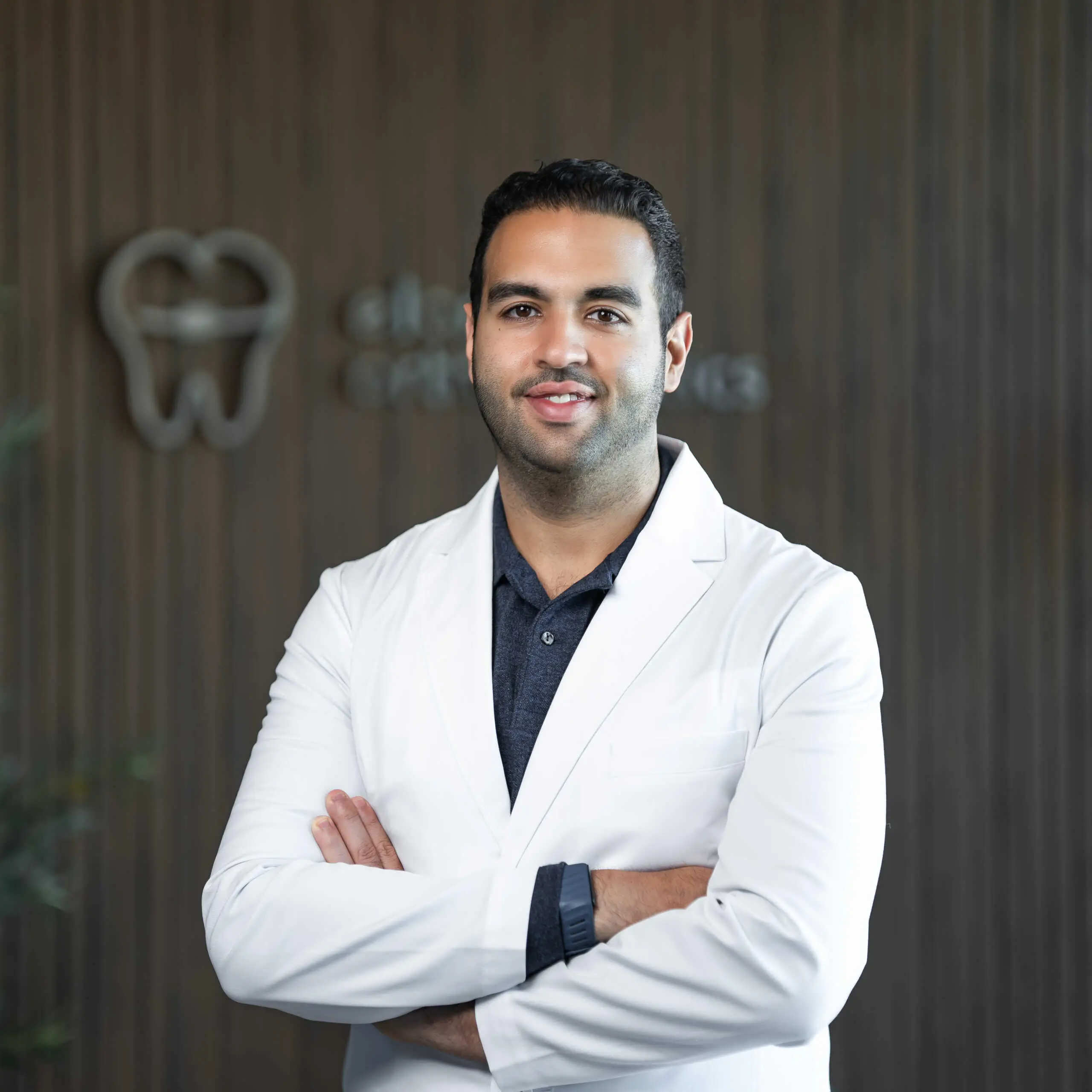 Dr. Sam Elkenawy: Orthodontist in Houston, TX Dr. Sam Elkenawy, orthodontic expert, in a white lab coat stands with arms crossed before a tooth symbol wall at Elara Orthodontics in Houston, TX.