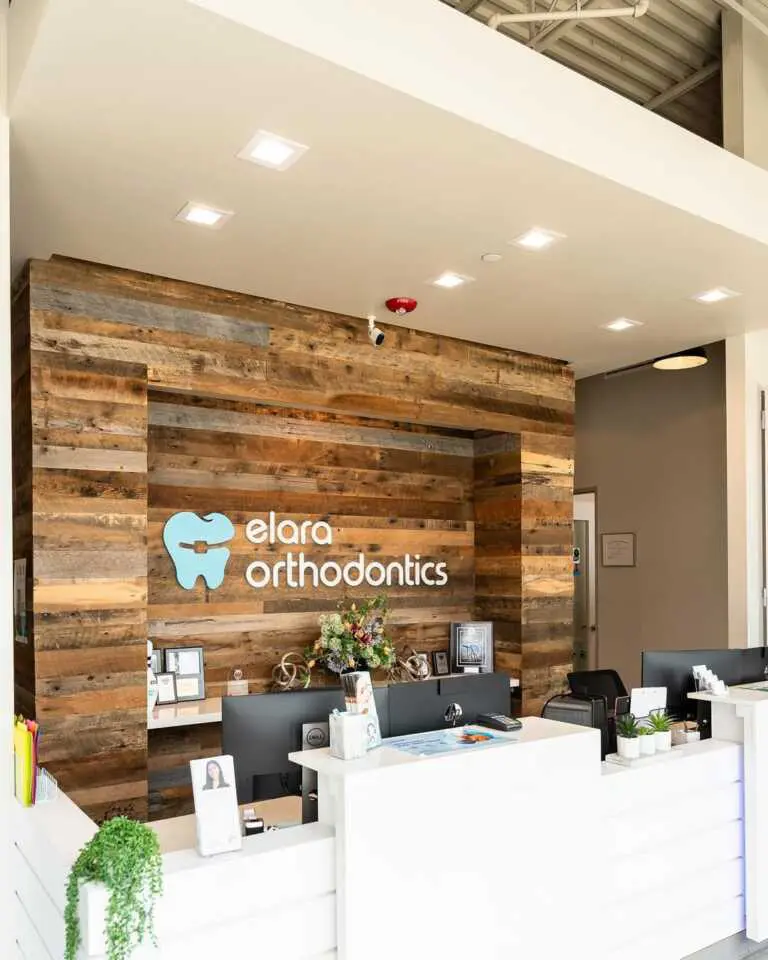 The reception area at Elara Orthodontics in Richmond or Houston, TX features a wood accent wall with the logo sign, fresh flowers, brochures, and computer stations.
