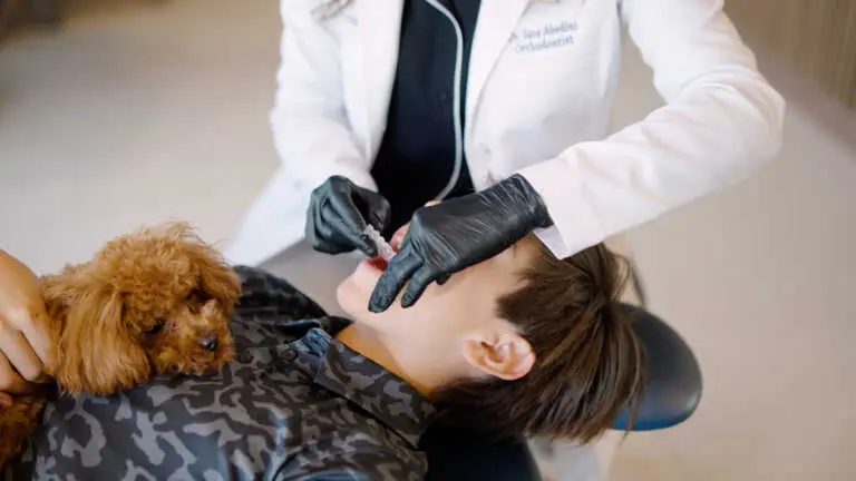 At Elara Orthodontics in Richmond or Houston, TX, a patient receives dental care while reclining in a chair and holding a small brown dog, attended by a dentist wearing black gloves and a white coat.