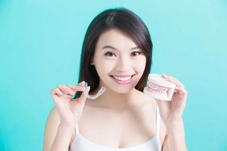 A woman holds a clear dental aligner and a dental model with braces, showcasing the Invisalign vs Braces cost in Richmond or Houston, TX comparison at Elara Orthodontics , set against a turquoise background.