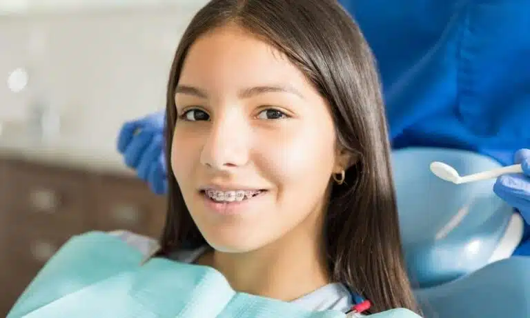 A teenage girl with long brown hair and braces smiles in a dental chair while a dental hygienist holds a tool nearby at Elara Orthodontics in Fulshear, TX.