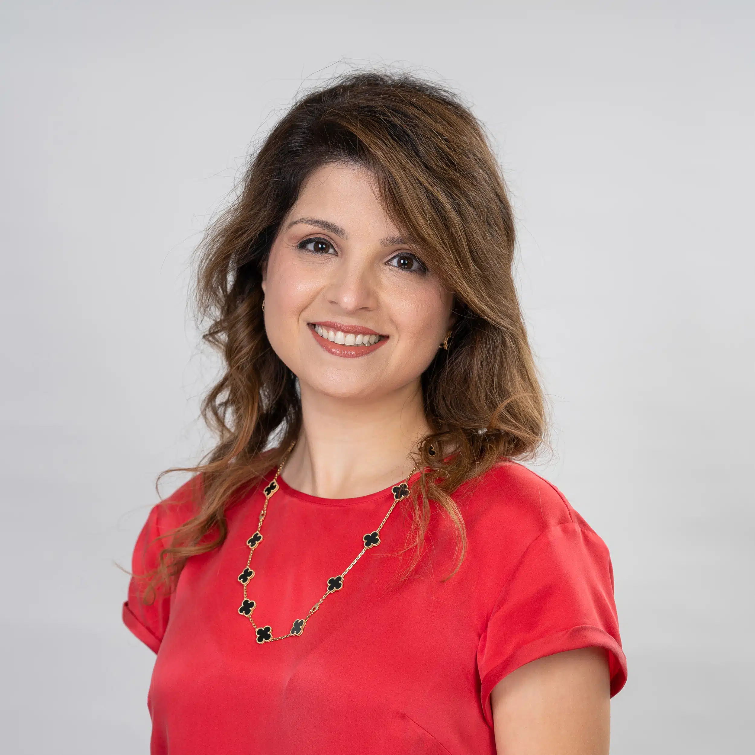 Dr-Sara Headshot of Dr. Sara Abedini, an orthodontist expert in Houston TX, smiling confidently against a white background. She has brown hair and wears a red top and a distinctive black clover necklace.