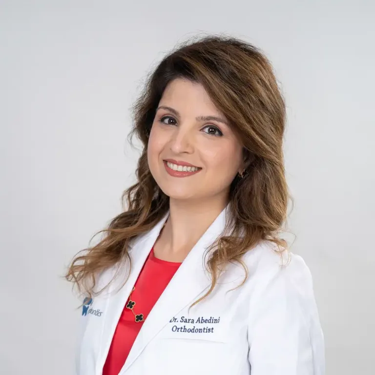 Dr. Sara Abedini: Orthodontist in Richmond and Houston, TX At Elara Orthodontics in Richmond or Houston, TX, Dr. Sara Abedini—recognized by her long brown hair, white lab coat, and red top—smiles warmly at the camera against a plain light background.