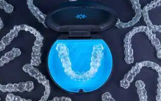Displayed on a dark surface, Invisalign clear dental aligners are shown in a stylish blue and black case, with several other aligners nearby—available from Elara Orthodontics in Richmond or Houston, TX.