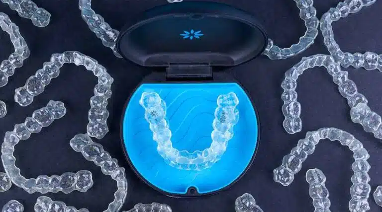 Displayed on a dark surface, Invisalign clear dental aligners are shown in a stylish blue and black case, with several other aligners nearby—available from Elara Orthodontics in Richmond or Houston, TX.