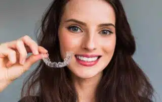 A woman with long brown hair smiles at the camera while holding an Invisalign clear dental aligner near her mouth, showcasing the service offered by Elara Orthodontics in Richmond or Houston, TX.