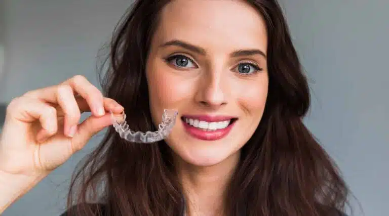 A woman with long brown hair smiles at the camera while holding an Invisalign clear dental aligner near her mouth, showcasing the service offered by Elara Orthodontics in Richmond or Houston, TX — a great example when considering Invisalign vs Braces.