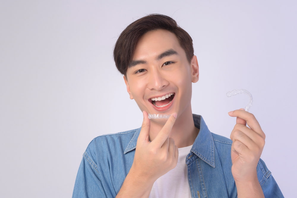 A person in a denim shirt smiles and points to an Invisalign clear dental aligner against a plain background, highlighting Elara Orthodontics in Richmond, TX.