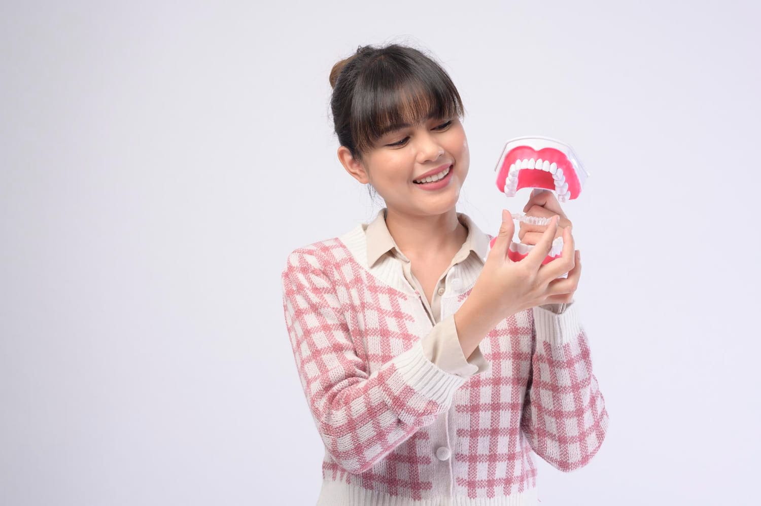 A woman in a checkered cardigan smiles while examining a model of human teeth, considering Invisalign treatment at Elara Orthodontics in Richmond, TX, against a plain white background.