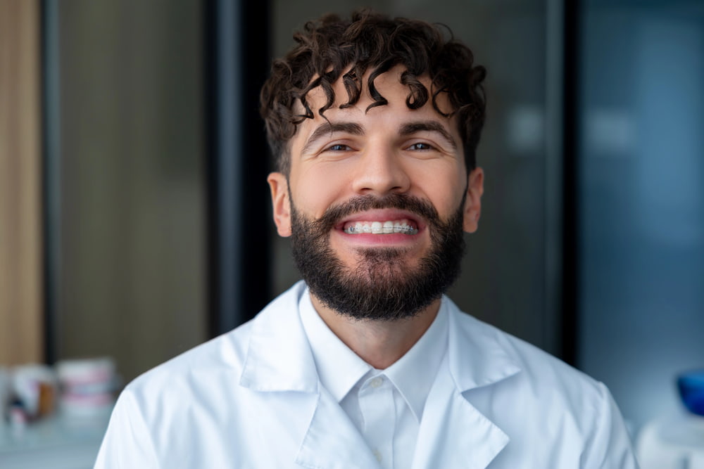 At Elara Orthodontics in Richmond or Houston, TX, a curly-haired man with a beard smiles at the camera while wearing a white lab coat indoors, representing Clear Braces treatment options.