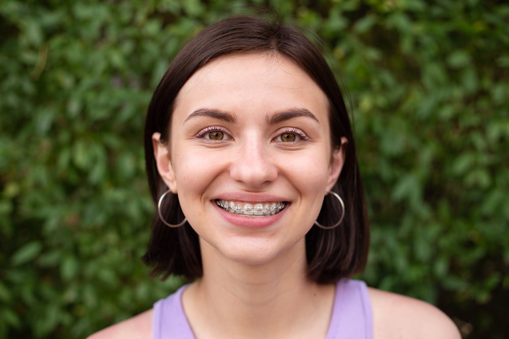 Smiling young woman with braces, hoop earrings, and purple top; greenery behind her for Elara Orthodontics in Richmond or Houston, TX.