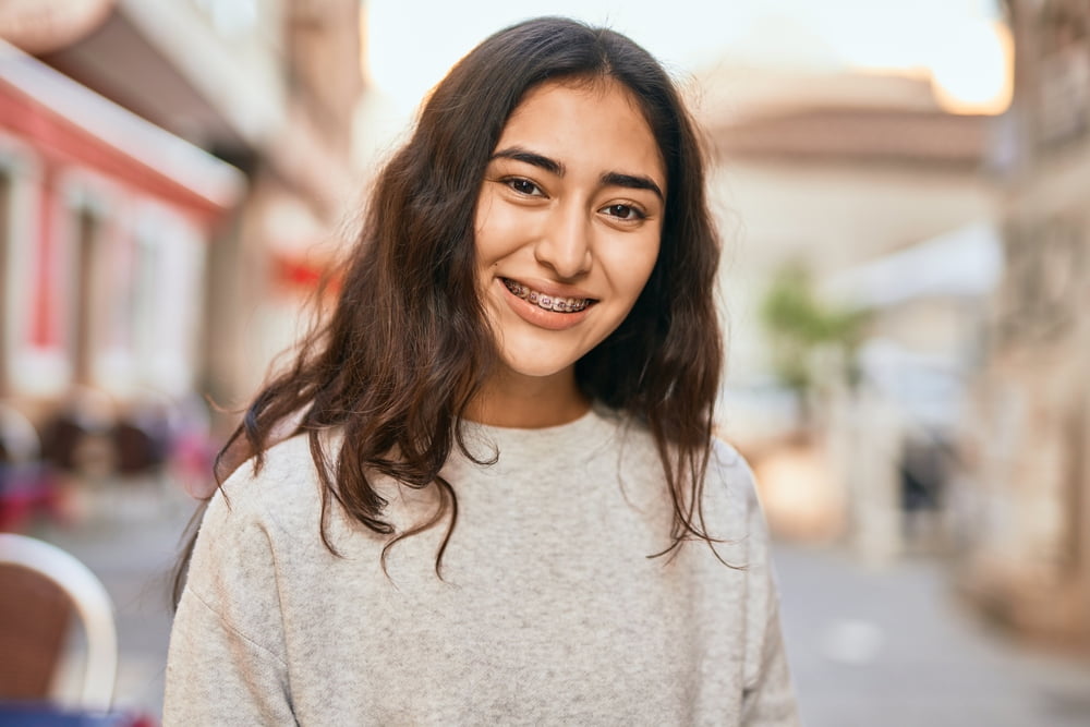Smiling with braces, a young person in a gray sweater stands outside Elara Orthodontics in Richmond, TX; cityscape behind.