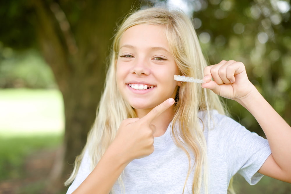 A smiling girl with long blonde hair holds Clear Aligners and points at them outdoors in front of trees, demonstrating benefits recommended by a children’s orthodontist at Elara Orthodontics in Richmond or Houston, TX.