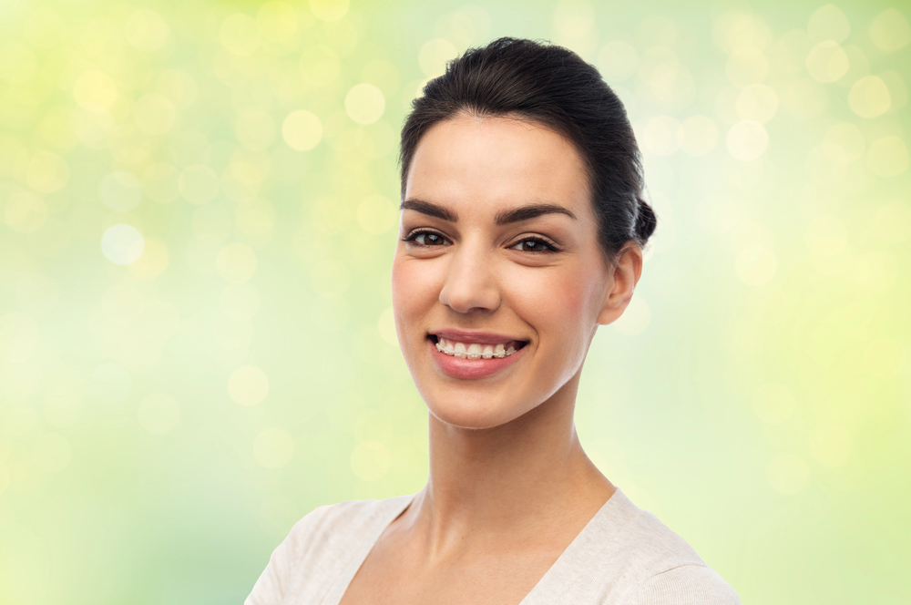 Smiling woman with dark hair and clear braces at Elara Orthodontics in Richmond, TX; white top, blurred green-yellow background.
