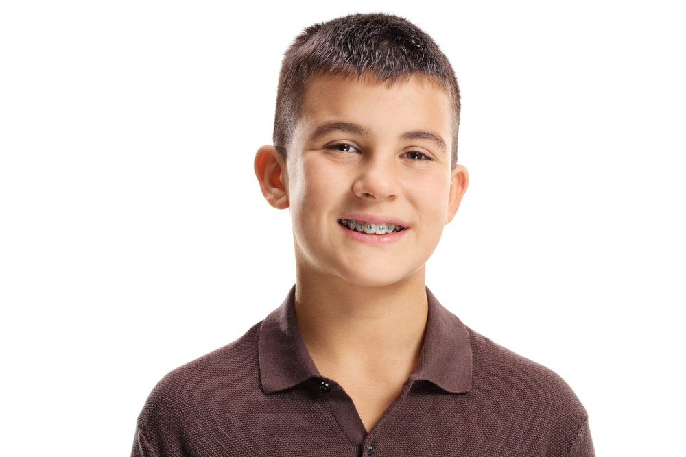 Smiling with traditional braces against a plain white background, a boy with short dark hair and a brown polo shirt showcases the results children can expect from Elara Orthodontics in Richmond or Houston, TX.