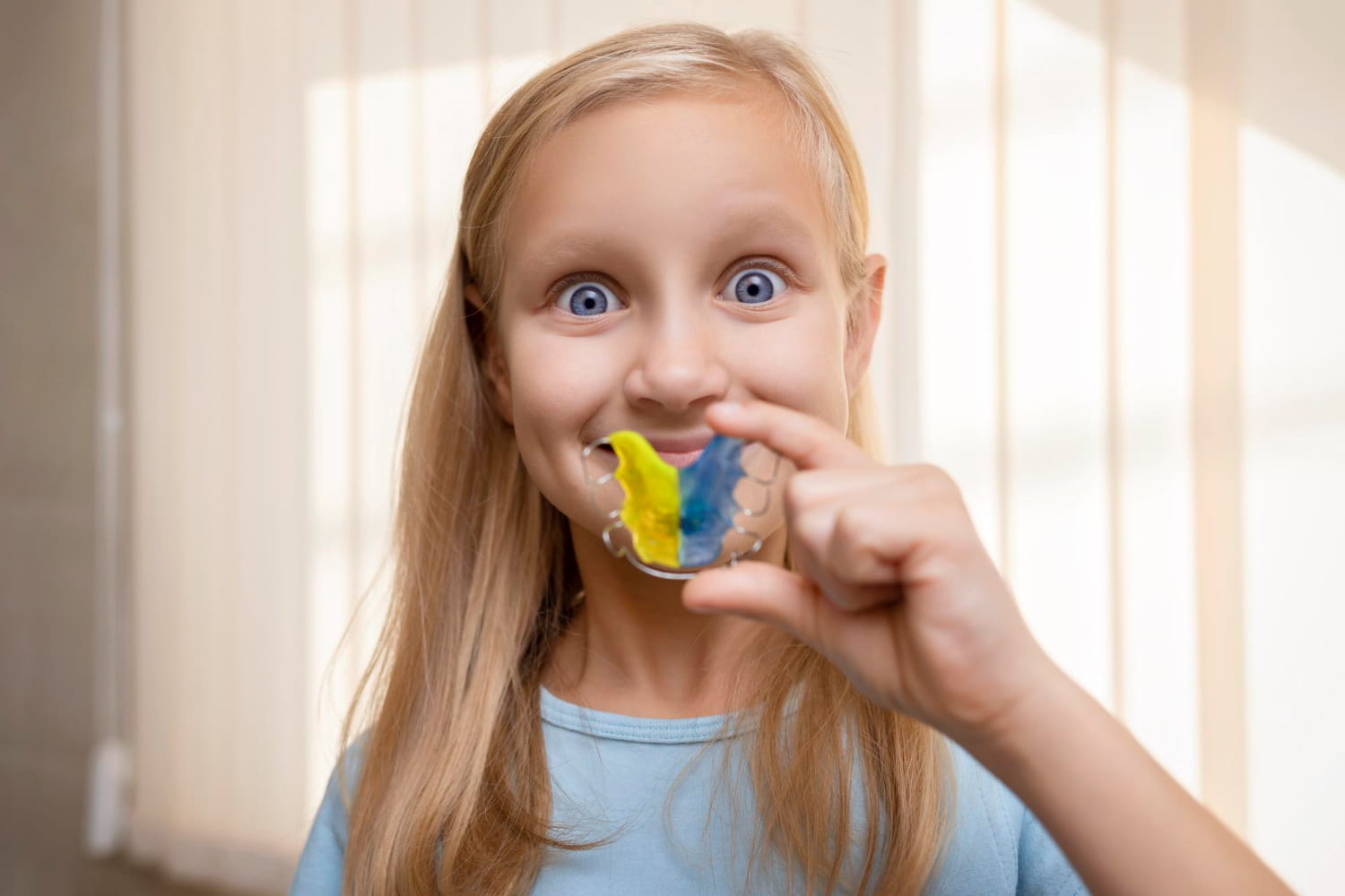 A young girl with long blonde hair smiles while holding a blue and yellow dental retainer in front of her mouth, highlighting the expertise of Elara Orthodontics in Richmond or Houston, TX.