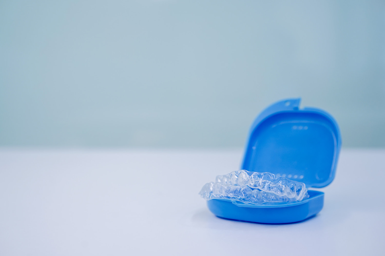 An Invisalign clear dental aligner from Elara Orthodontics rests inside an open blue case on a white surface, set against a plain light blue background in Richmond, TX.