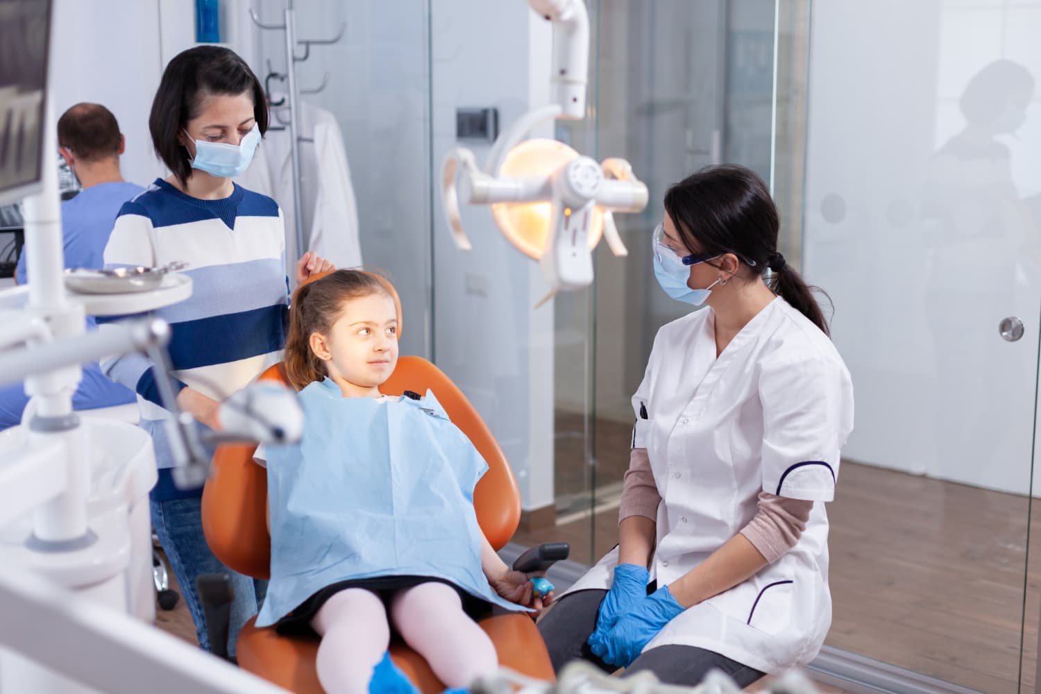In Richmond or Houston, TX, at Elara Orthodontics, a young girl sits in a dental chair wearing a bib as a masked dentist—possibly a children’s orthodontist—and an adult stand nearby in the clinic.