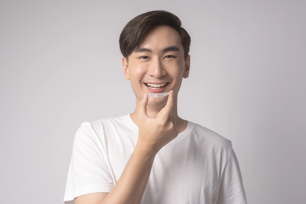A smiling person, possibly of Asian descent, holds up a clear Invisalign aligner against a light gray background. This image could relate to Invisalign options in Richmond, TX.