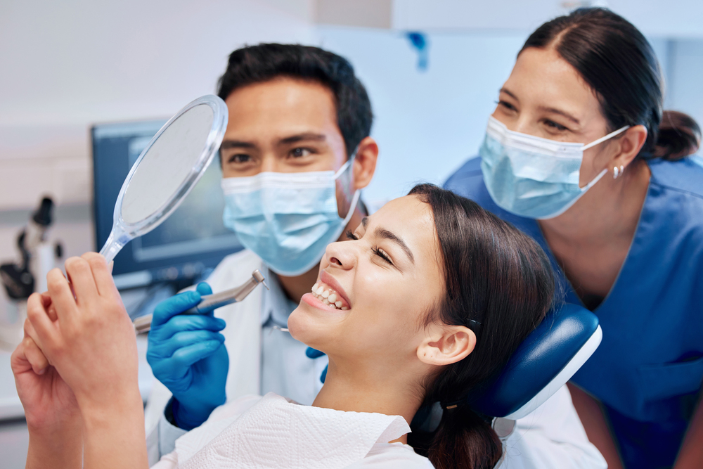 A smiling female patient looks at her teeth in a mirror, seemingly pleased, while a male orthodontist and a female dental assistant, both wearing masks, observe in a dental office in Richmond, TX.