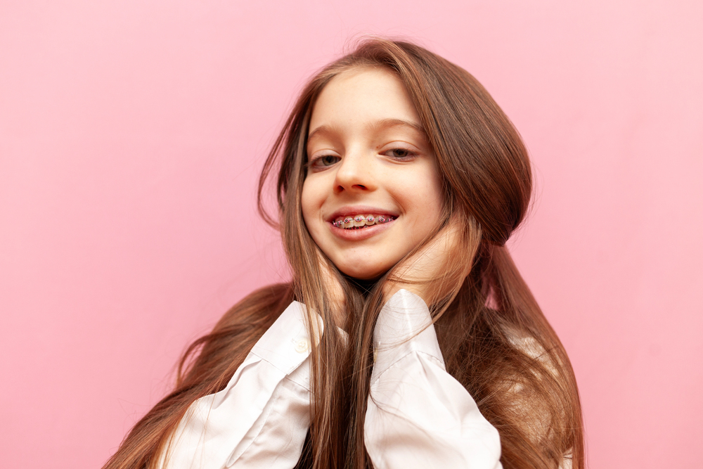A young girl with long brown hair and braces smiles charmingly, hands near her face, against a pink background. She represents a typical patient seeking an orthodontist in Richmond, TX.