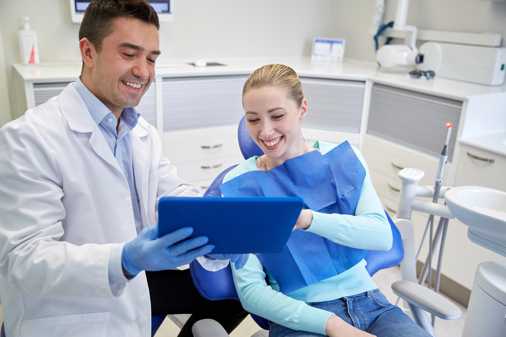 A male orthodontist in Richmond TX, in a white lab coat and blue gloves, shows a tablet to a smiling female patient seated in a dental chair with a bib.