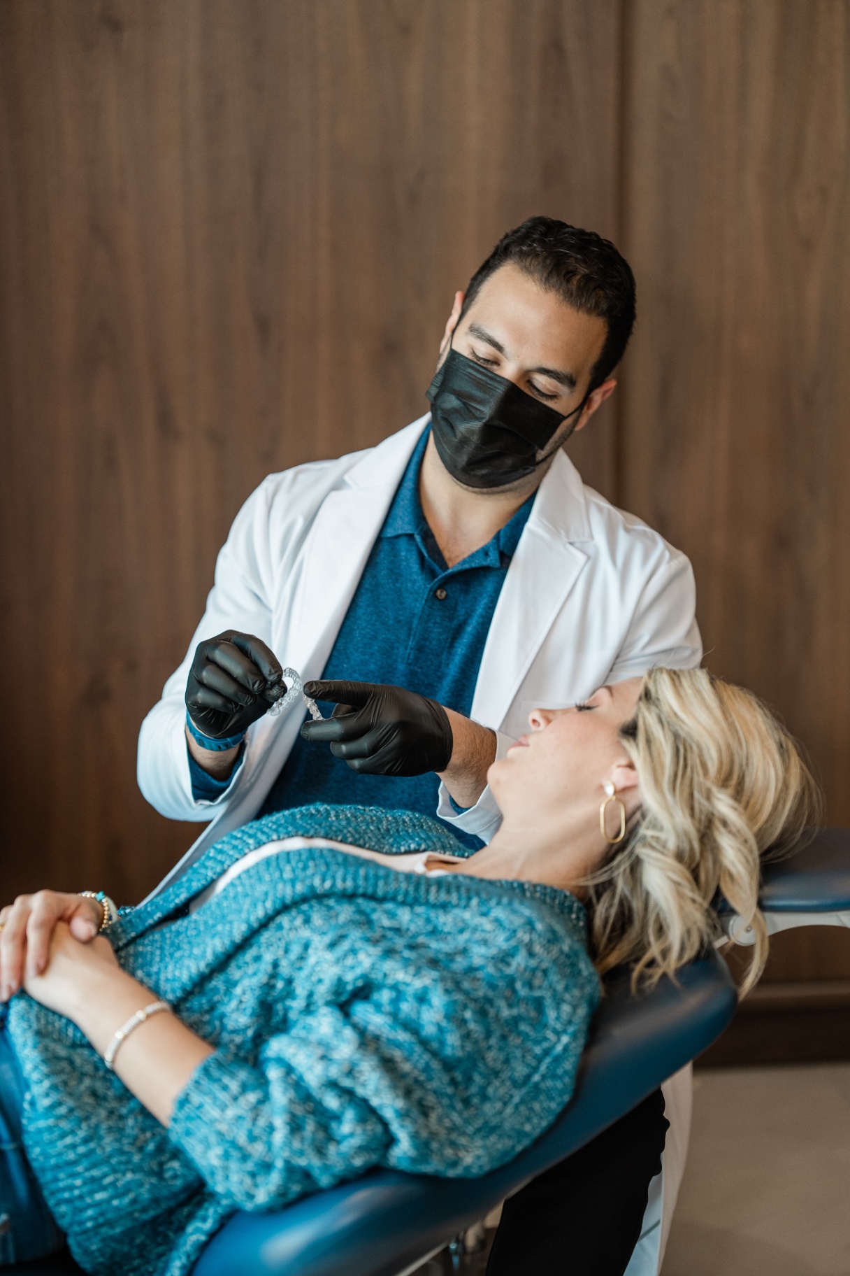 Dr. Sara Abedini and Dr. Sam Elkenawy: Orthodontist in Richmond TX At Elara Orthodontics in Richmond or Houston, TX, a dentist wearing a mask and gloves demonstrates a dental device to a female patient reclining in the dental chair.