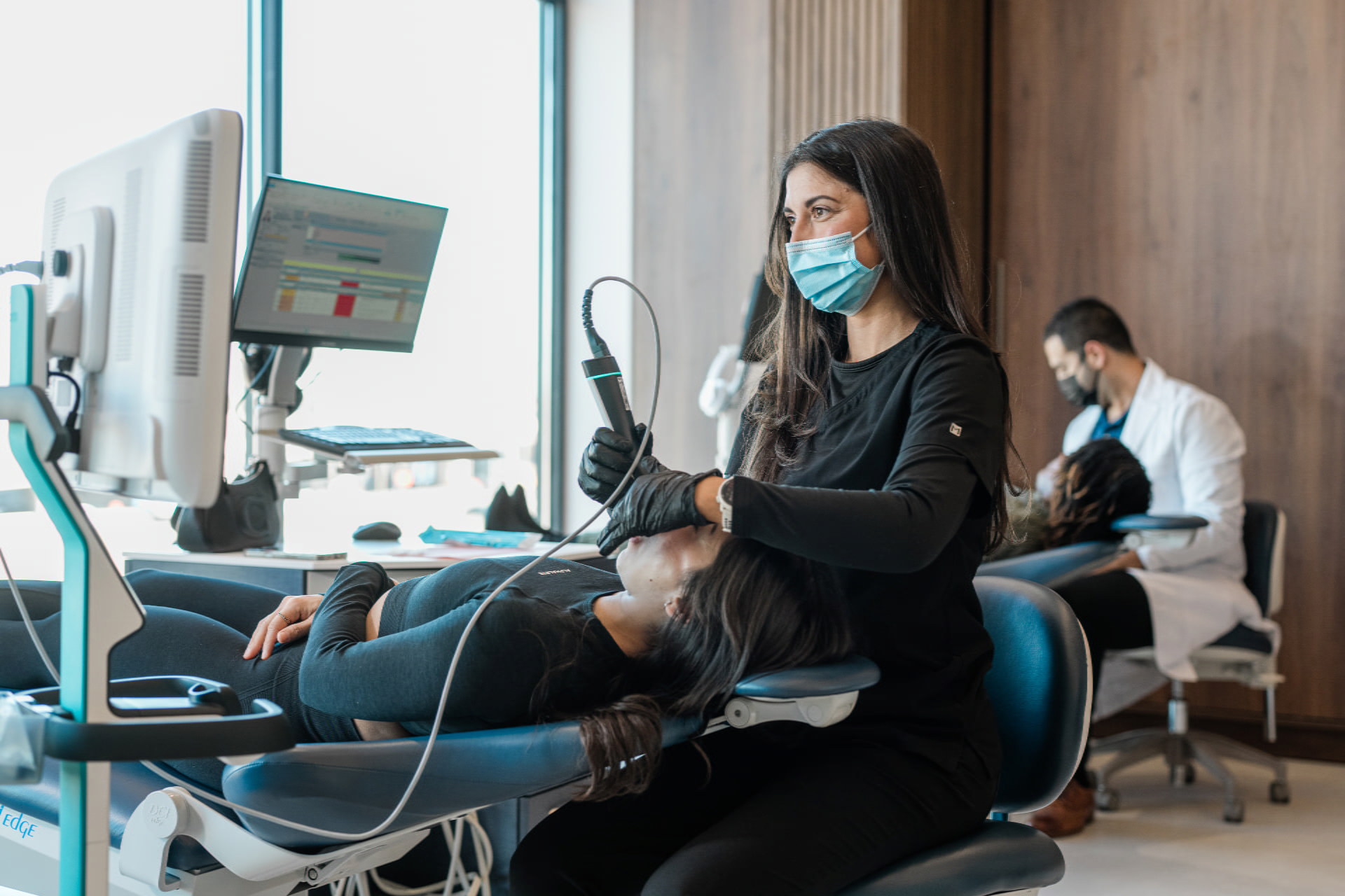 Dr. Sara Abedini and Dr. Sam Elkenawy: Orthodontist in Houston TX At Elara Orthodontics in Richmond or Houston, TX, a dental professional wearing a mask and gloves uses a digital scanner on a patient, with staff and other patients visible in the background.