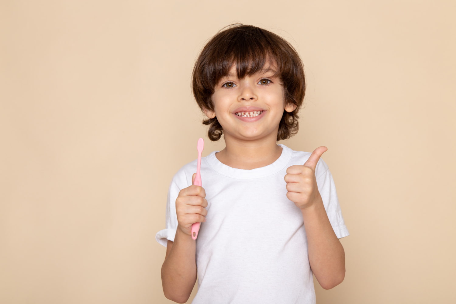 A smiling child with braces stands against a beige background, holding a pink toothbrush and giving a thumbs up after visiting Elara Orthodontics, a trusted Children’s Orthodontist in Richmond or Houston, TX.