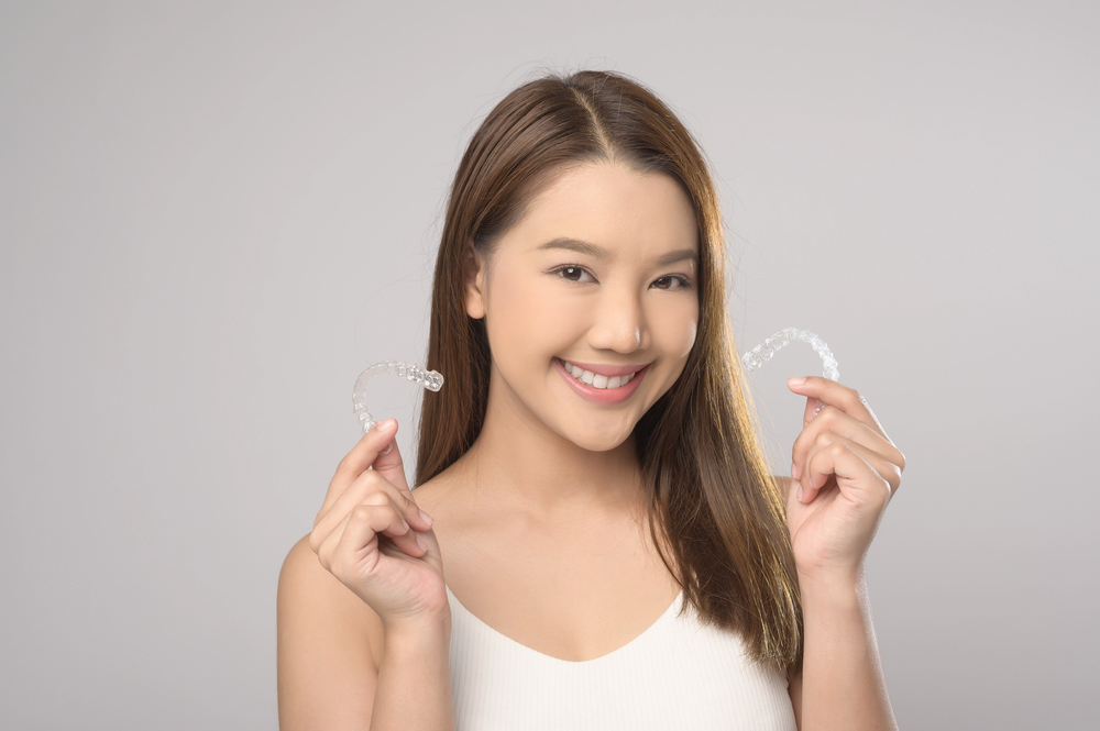 Smiling woman with long brown hair holds two Invisalign® Clear Aligners at Elara Orthodontics in Richmond, TX.