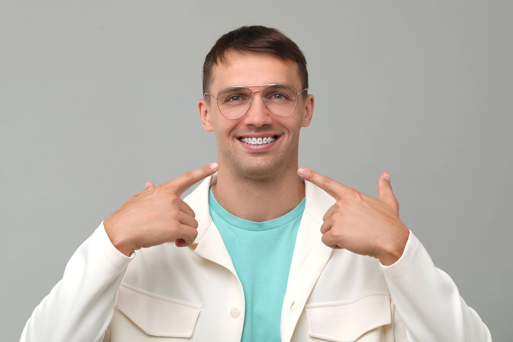 Smiling man in glasses and a white jacket points to metal braces, promoting Elara Orthodontics in Houston, TX.