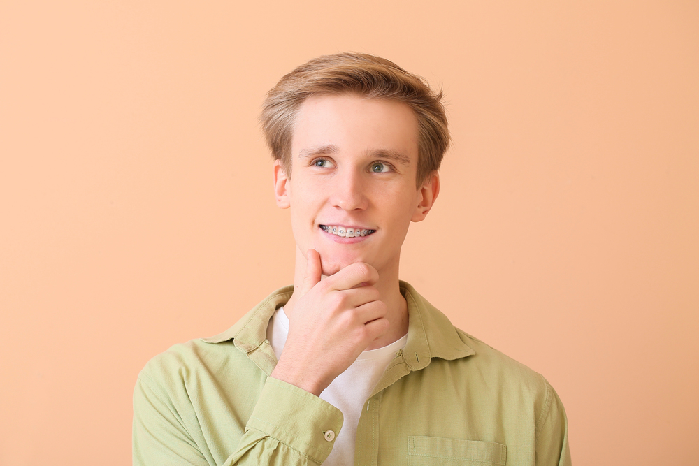 A smiling young man with metal braces at Elara Orthodontics in Richmond TX, wearing light green, looks up thoughtfully.