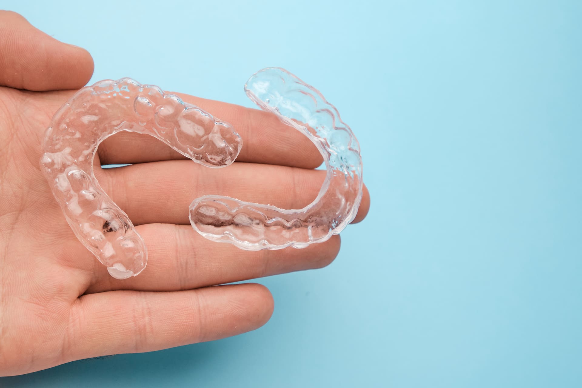 A hand displays two clear Invisalign aligners against a light blue background, showcasing options available at Elara Orthodontics in Houston, TX.