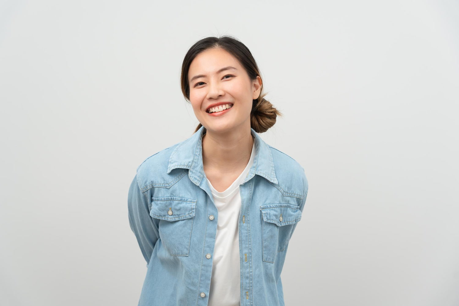 A confident woman with dark hair tied back smiles, showing her Invisalign aligners while wearing a light blue denim shirt over a white top against a plain background, representing Elara Orthodontics in Richmond, TX.