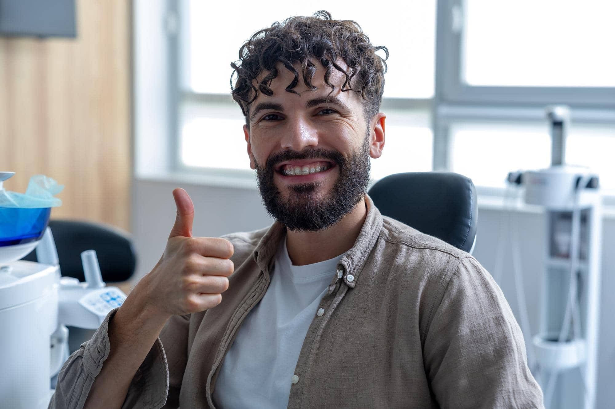 A man with curly hair and a beard smiles indoors, giving a thumbs up to show Elara Orthodontics clear ceramic braces in Houston, TX.