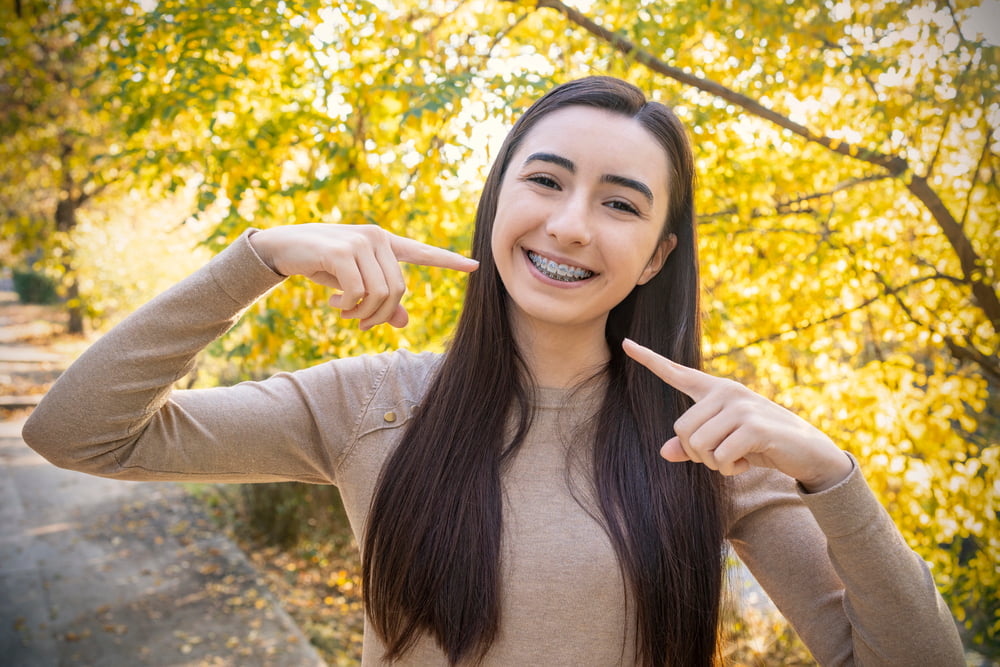 Smiling young woman with braces points to her teeth outdoors; schedule a free Elara Orthodontics consult in Richmond, TX.