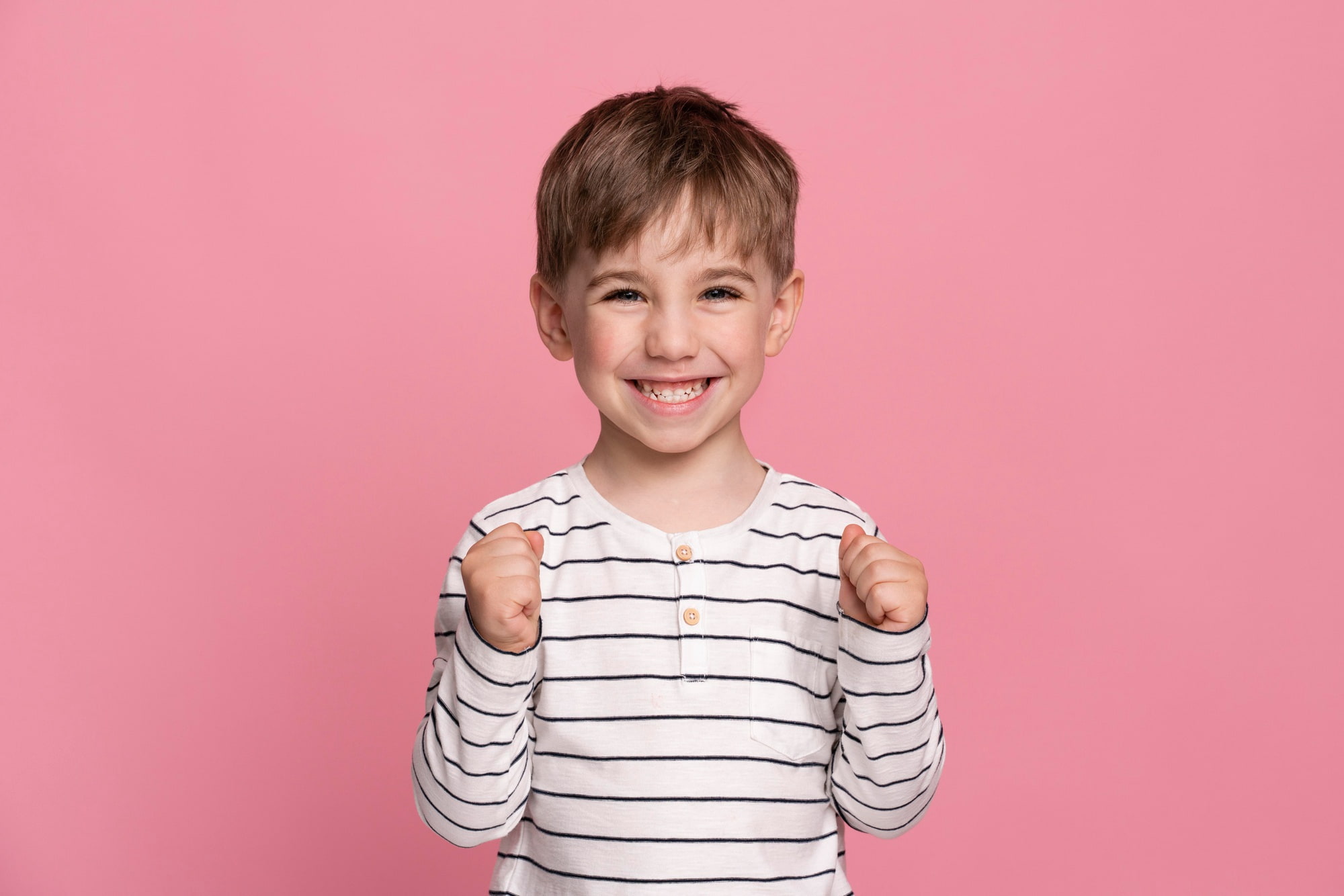 A young boy in a white and black striped shirt clenches his fists in excitement and smiles brightly, showing off his teeth—likely after a great visit to the Children’s Orthodontist at Elara Orthodontics in Richmond or Houston, TX.