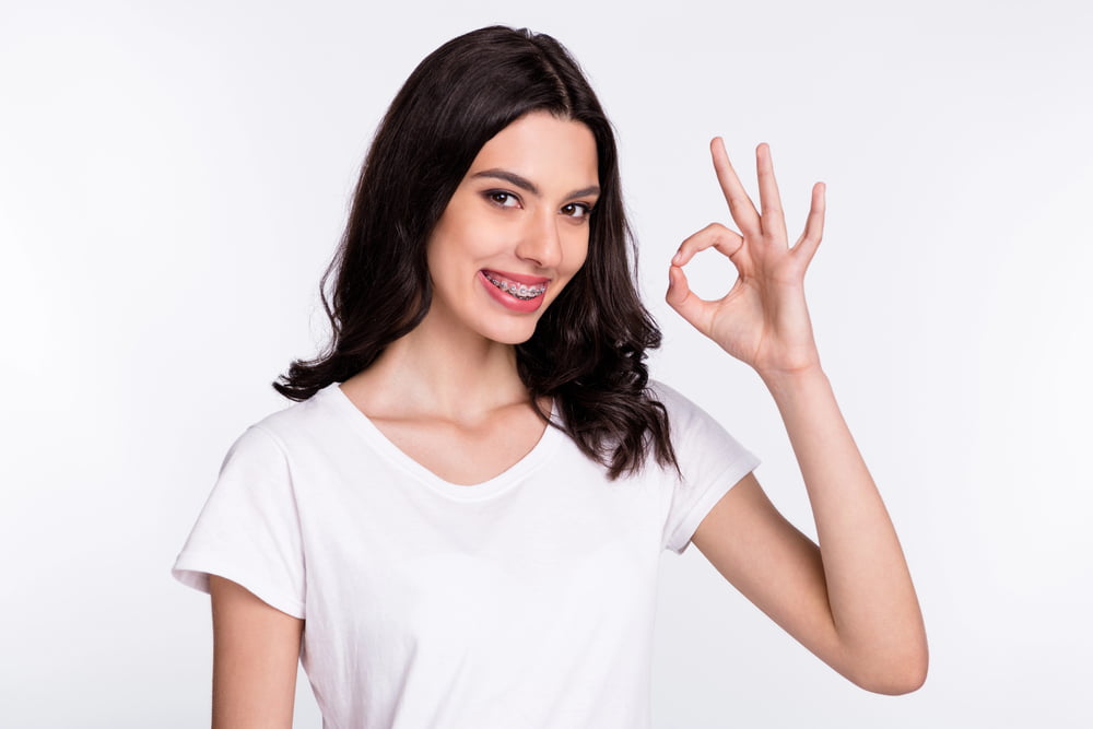 Smiling young woman with braces at Elara Orthodontics in Houston TX, wearing a white t-shirt and making an "OK" gesture.