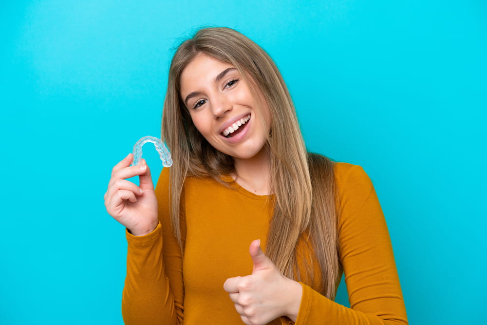 A young woman with long hair, wearing a mustard shirt, stands against a bright blue background holding an Invisalign clear aligner and giving a thumbs up—representing Elara Orthodontics in Houston, TX.