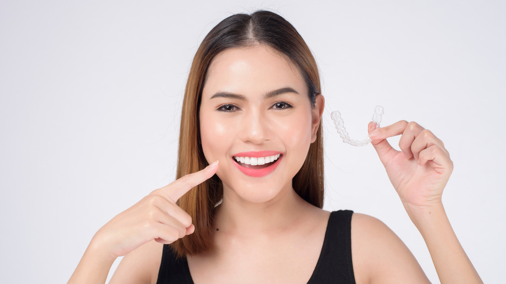 A woman smiles and points to her teeth while holding an Invisalign clear aligner, showcasing Elara Orthodontics’ services in Richmond, TX, against a plain white background.