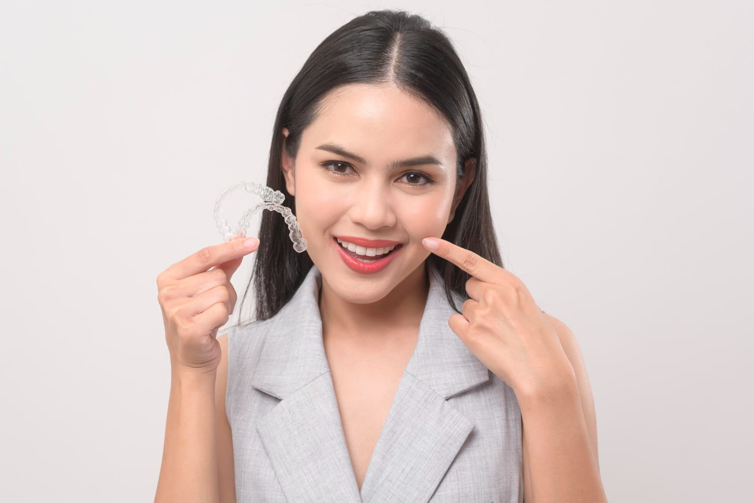 A woman wearing a light gray sleeveless top smiles and points at her teeth while holding an Invisalign clear aligner, representing Elara Orthodontics in Houston, TX, against a plain background.