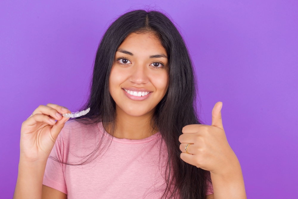 brunette girl wearing pink shirt purple background holding Invisalign clear aligners - how long does Invisalign take in Richmond and Houston, TX.