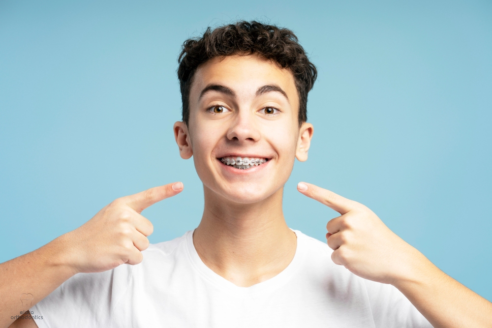 A teenage boy with curly hair in a white t-shirt smiles and points to his braces against a blue background, showcasing Elara Orthodontics as your source for Invisalign vs Braces cost in Richmond or Houston, TX.