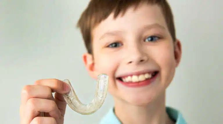 A smiling child displays a clear dental mouthguard, highlighting Invisalign for Kids offered by Elara Orthodontics in Richmond or Houston, TX.