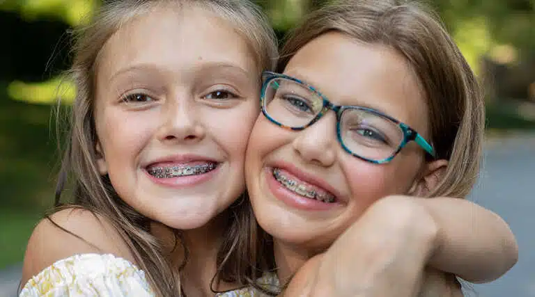 Smiling and hugging outdoors, two happy girls with Kids Braces—one in glasses—show off their smiles at Elara Orthodontics in Richmond or Houston, TX.