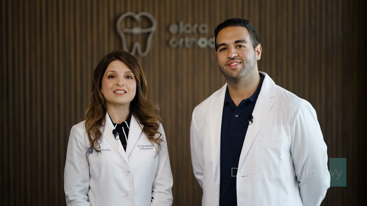 At Elara Orthodontics in Richmond or Houston, TX, Dr. Sara Abedini and Dr. Sam Elkenawy—both in white coats—pose before a wooden wall displaying a tooth logo and the clinic’s name.